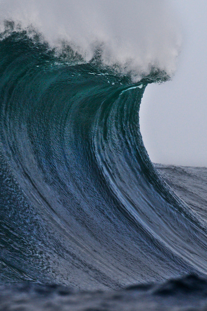 Large ocean wave face topped with white foam against a gray sky